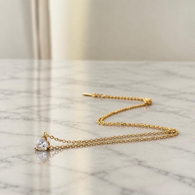 Collana Punto Luce Cuore Oro con catena dorata in acciaio inossidabile e pendente a cuore trasparente sfaccettato su marmo bianco.